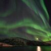 Aurores boréales vertes déployées au-dessus d’un fjord norvégien, avec la lune qui se reflète dans l’eau