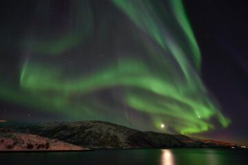 Aurores boréales vertes déployées au-dessus d’un fjord norvégien, avec la lune qui se reflète dans l’eau