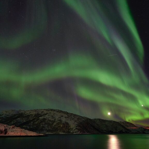 Aurores boréales vertes déployées au-dessus d’un fjord norvégien, avec la lune qui se reflète dans l’eau