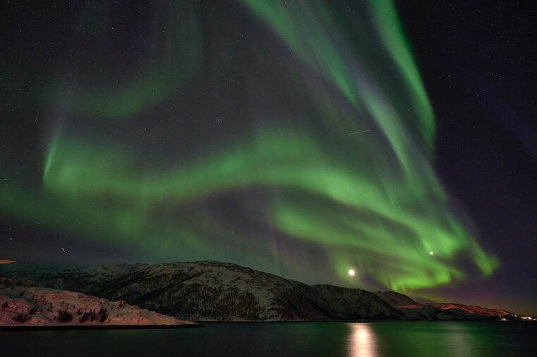 Aurores boréales vertes déployées au-dessus d’un fjord norvégien, avec la lune qui se reflète dans l’eau