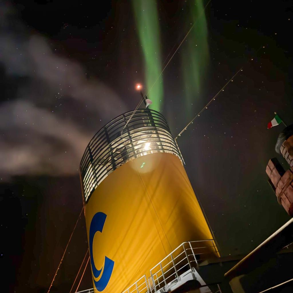La cheminée jaune ornée du C bleu de Costa, éclairée de nuit, avec des rubans d’aurore boréale verte dans le ciel étoilé