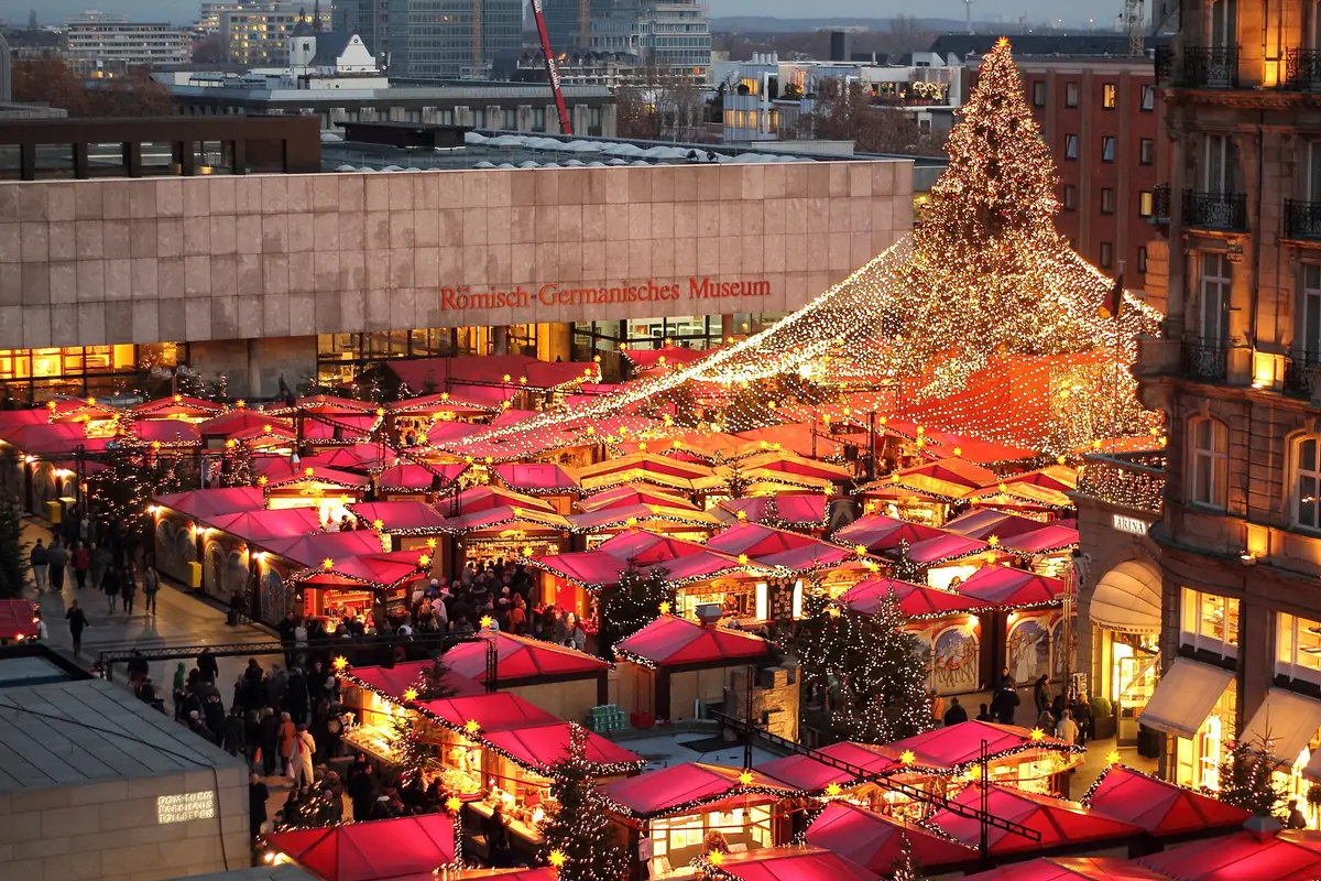 Vue d’ensemble du marché de Noël de Cologne au crépuscule : chalets rouges, grand sapin lumineux et cathédrale