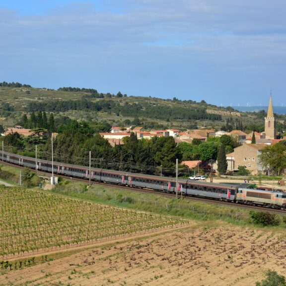 Train Intercités Bordeaux-Marseille tracté par une locomotive grise et orange, entre les vignes et le village de Névian, près de Narbonne
