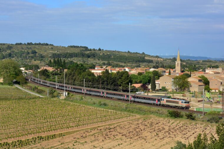 Train Intercités Bordeaux-Marseille tracté par une locomotive grise et orange, entre les vignes et le village de Névian, près de Narbonne