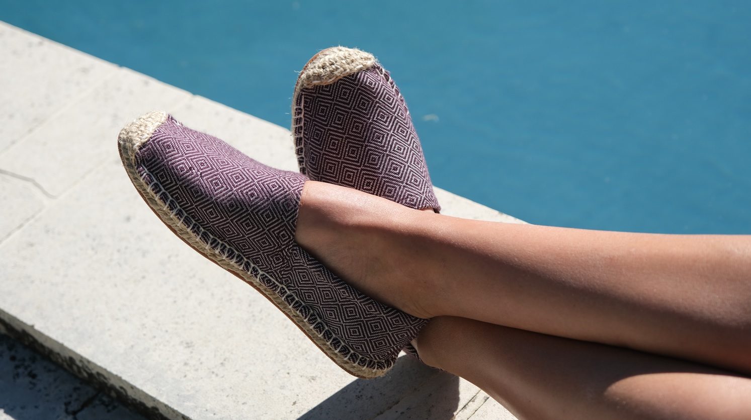 Pieds croisés au bord d'une piscine, chaussés d'espadrilles en tissu de krama prune à semelle de jute tressé.