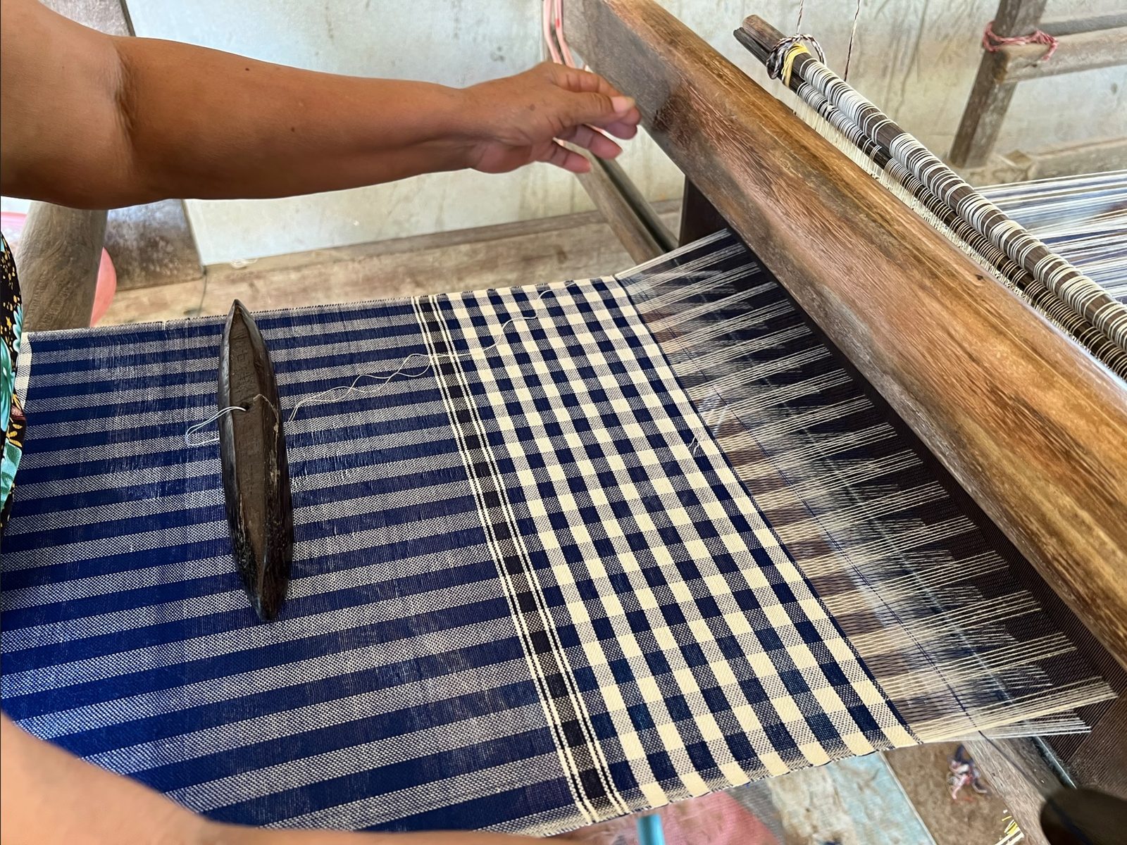 Main d'une tisserande sur un métier à tisser en bois, un krama à carreaux bleu marine et blanc en cours de tissage.