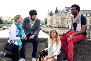 Quatre amis assis sur les quais de Seine face à la Conciergerie, écharpes Krama Heritage à carreaux et à rayures autour du cou.