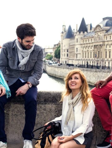 Quatre amis assis sur les quais de Seine face à la Conciergerie, écharpes Krama Heritage à carreaux et à rayures autour du cou.