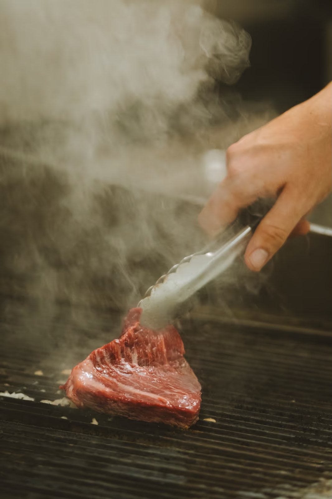 Pièce de contre-filet saisie à la pince sur un grill, dans un nuage de fumée.