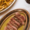 Contre-filet tranché nappé de sauce dans un plat ovale en métal, à côté d'un plat de frites et d'un verre de vin rouge.