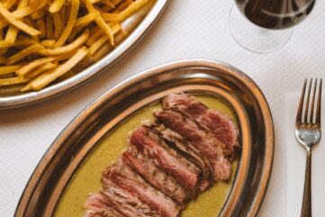 Contre-filet tranché nappé de sauce dans un plat ovale en métal, à côté d'un plat de frites et d'un verre de vin rouge.