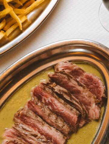 Contre-filet tranché nappé de sauce dans un plat ovale en métal, à côté d'un plat de frites et d'un verre de vin rouge.